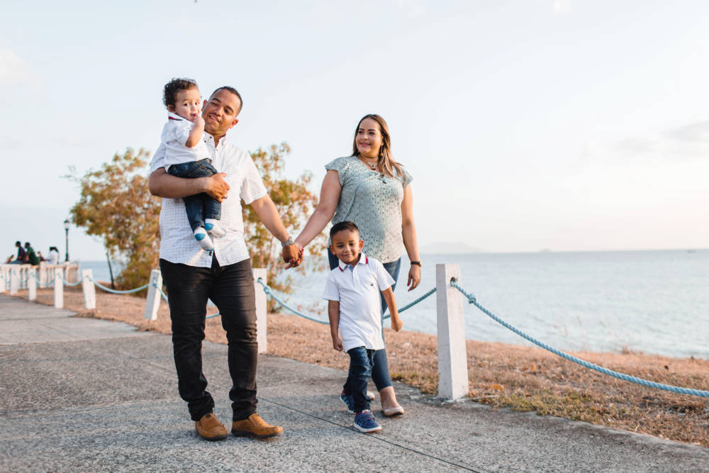Aseguranza Medica Barata - Seguro Medico en Estados Unidos - Familia Latina caminando en el parque madre y padre agarrados de manos y cargando a hijo